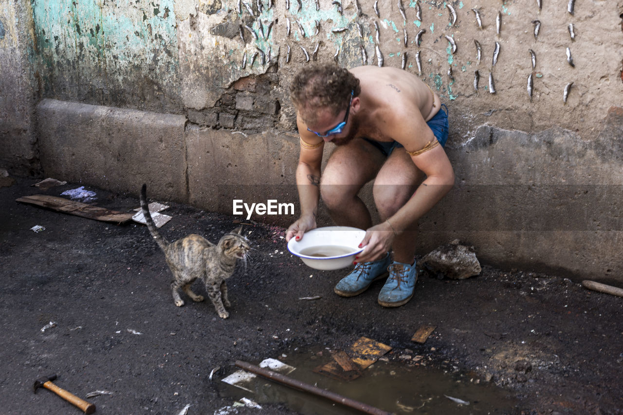 Full length of a shirtless man giving water to a cat against a wall