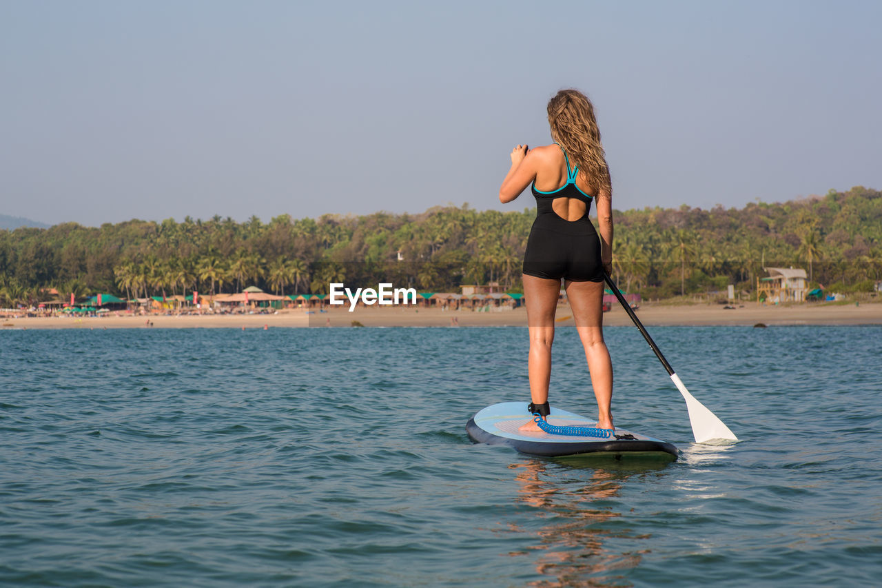 Rear view of mid adult woman paddleboarding on sea against clear sky