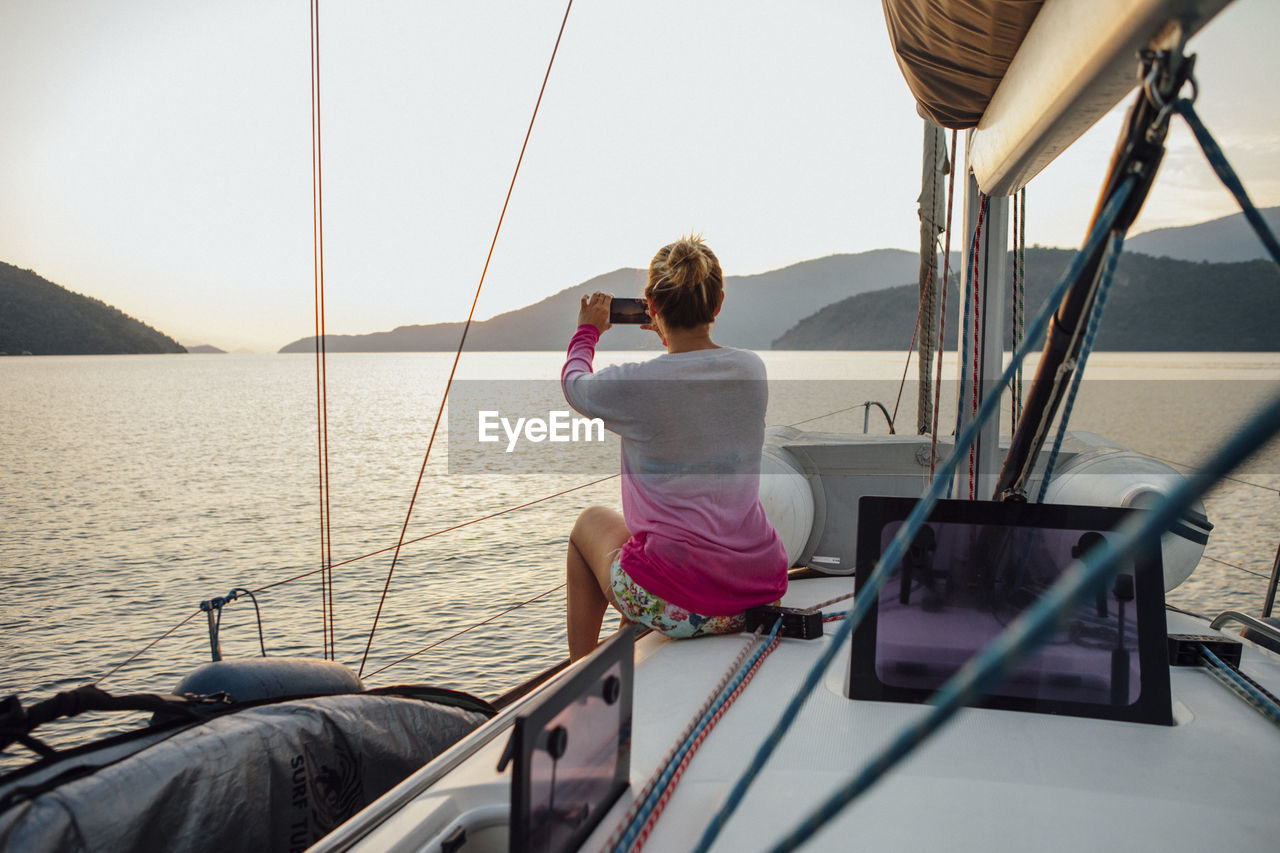 Rear view of woman photographing while traveling in sailboat on sea against clear sky