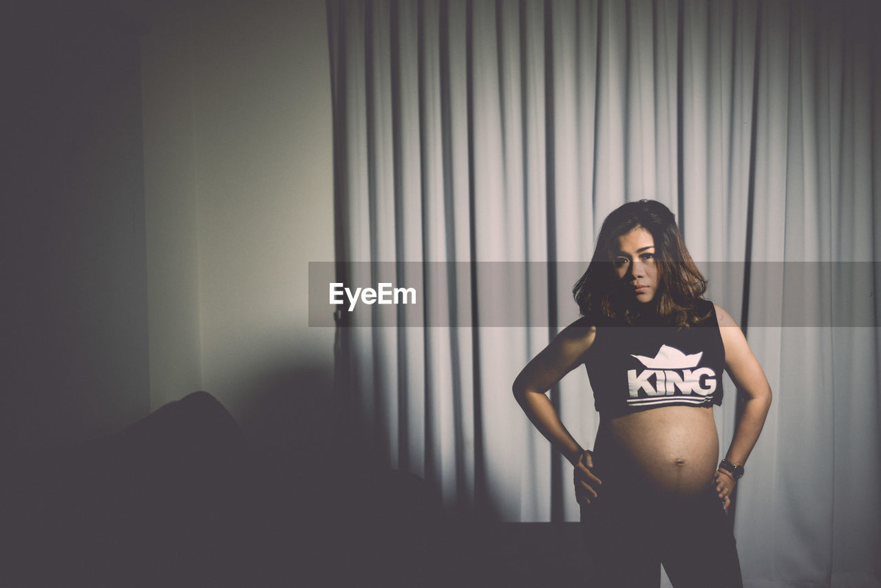 Portrait of pregnant woman standing against curtains at home