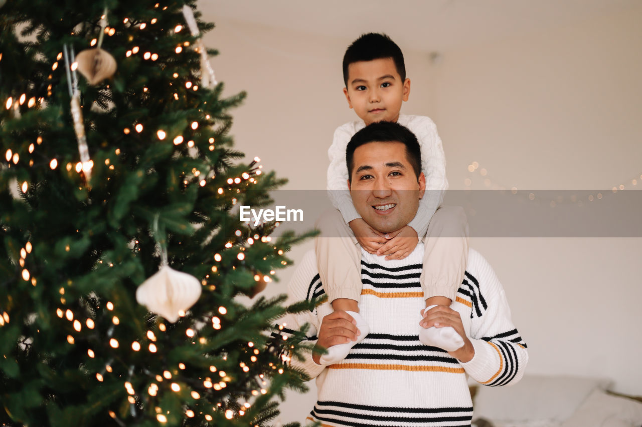 Chinese multiracial dad and little son in sweaters decorate the christmas tree at home on holiday