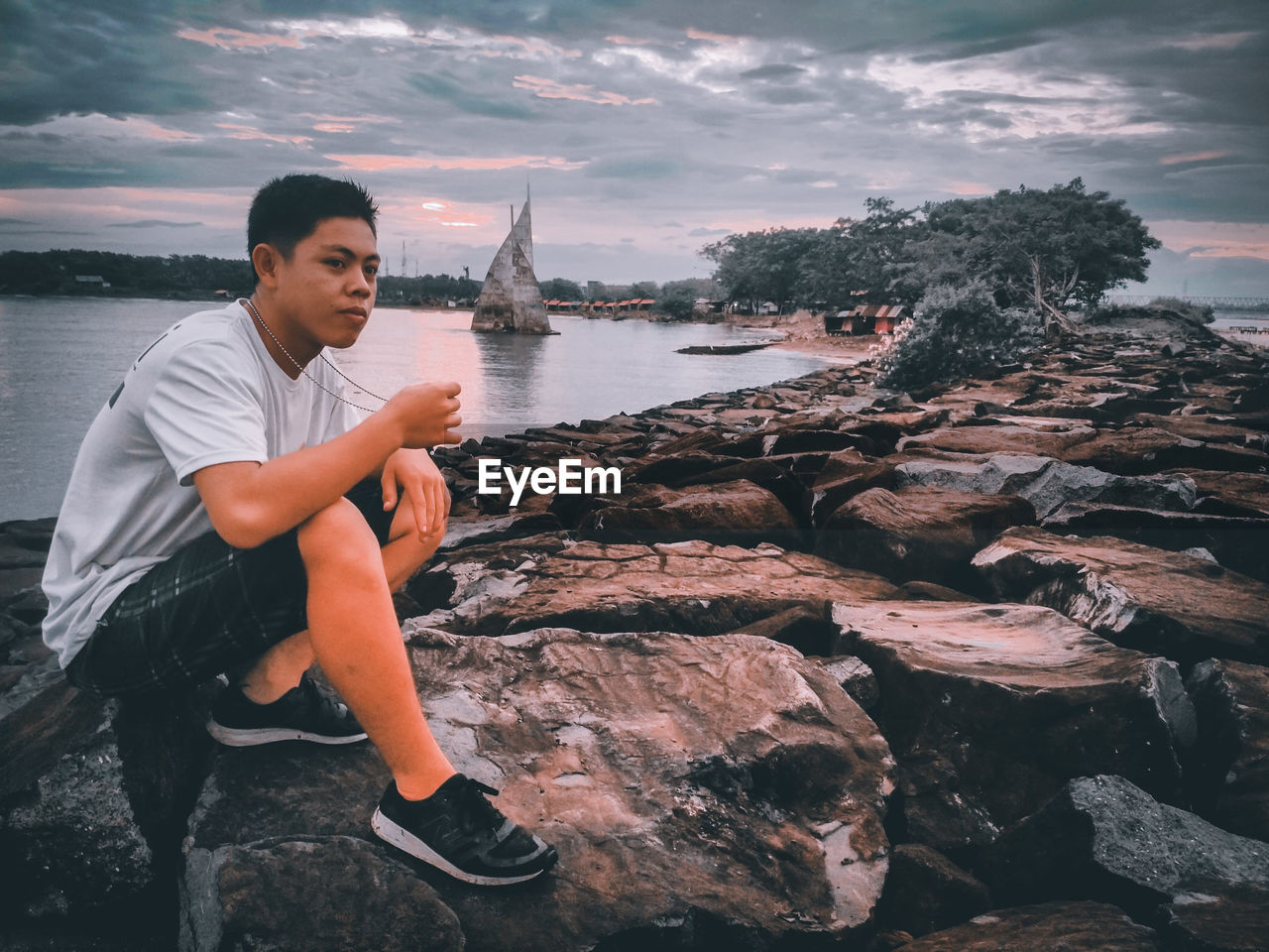 Young man sitting on rock by sea against sky