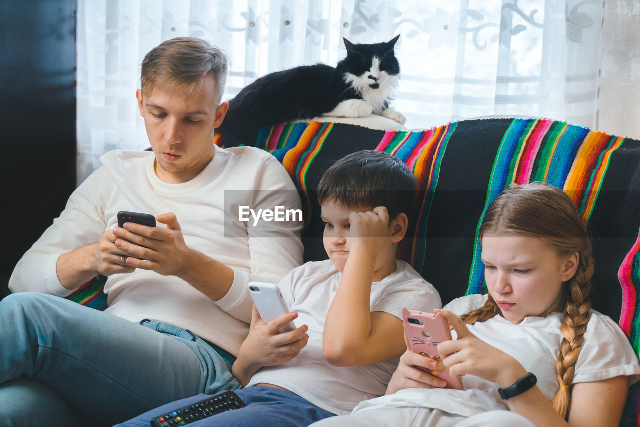 Siblings using smart phones while sitting on sofa with cat at home