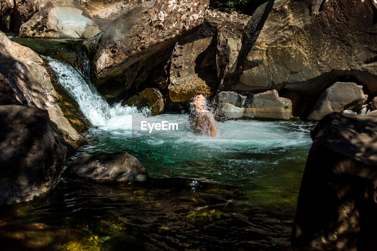 Male tourist emerging from clean pond water near cascade on sunny day in mountains