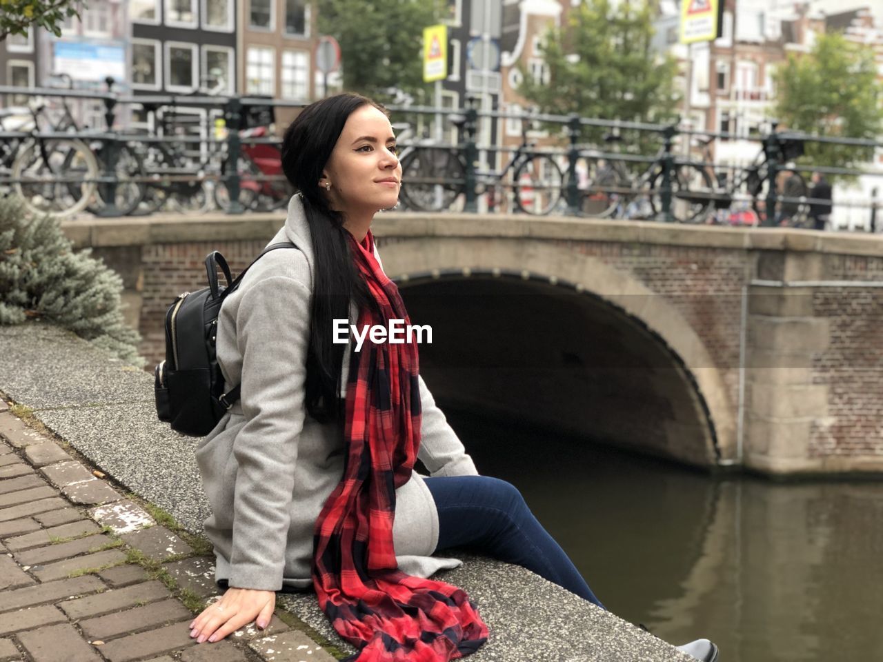 Young woman looking away while sitting by river in city