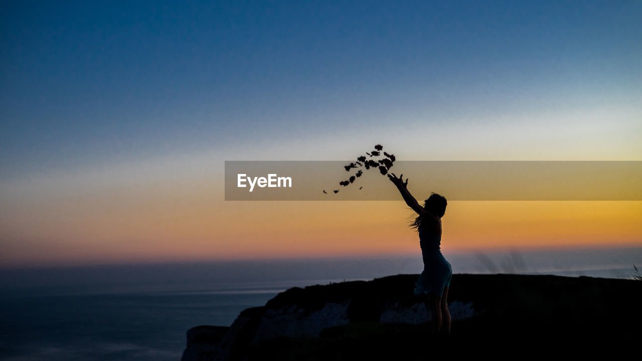 Silhouette of woman throwing leaves against sky during sunset