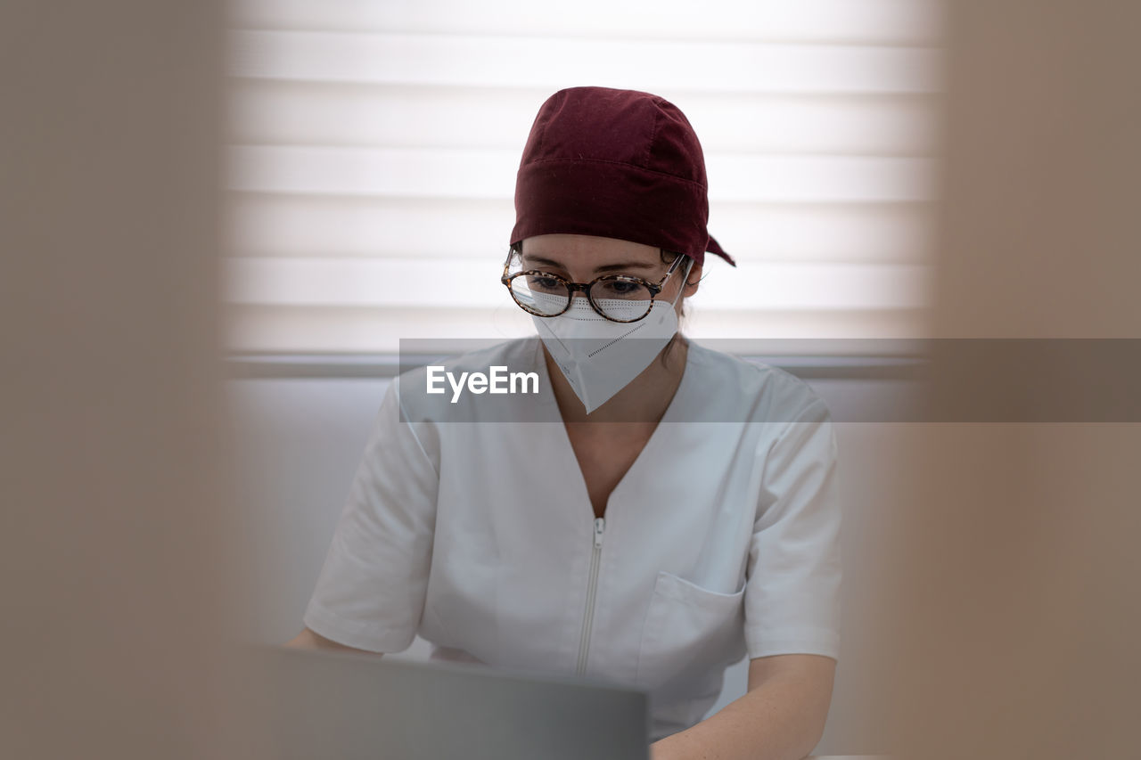 Concentrated female doctor in medical mask and uniform sitting at table and browsing laptop while working in modern workspace