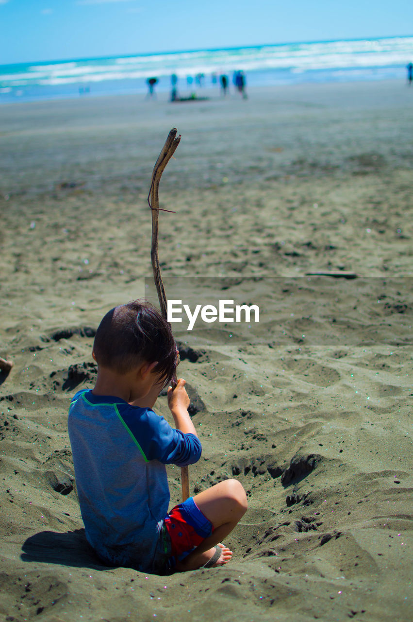 Boy with stick sitting at beach on sunny day