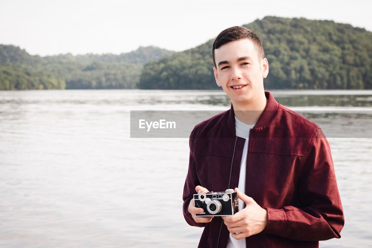 Portrait of young man holding camera against lake and sky