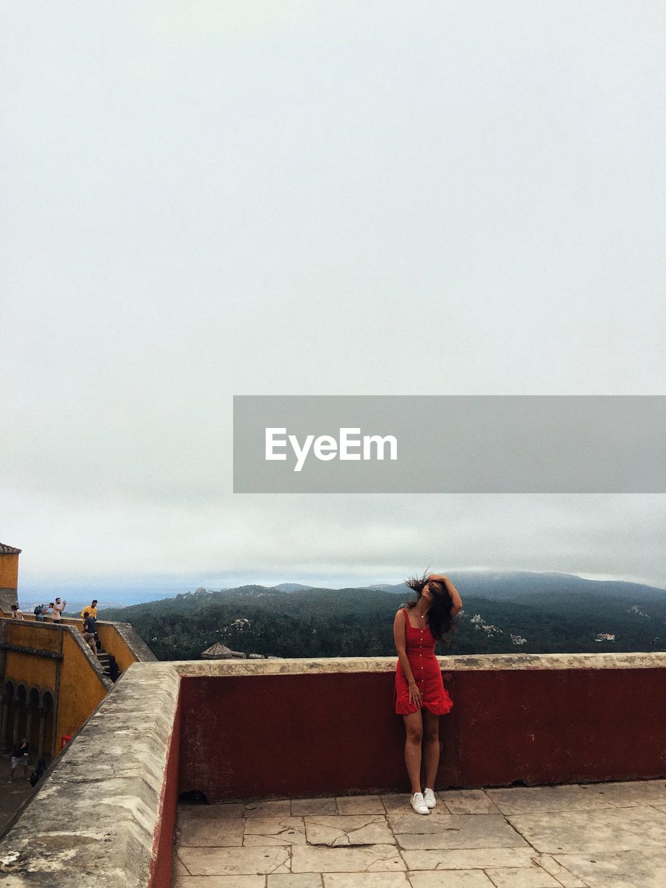 FULL LENGTH REAR VIEW OF WOMAN STANDING ON WALL AGAINST SKY