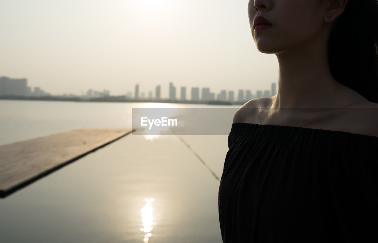 MIDSECTION OF WOMAN STANDING IN SEA AGAINST SKY DURING SUNSET