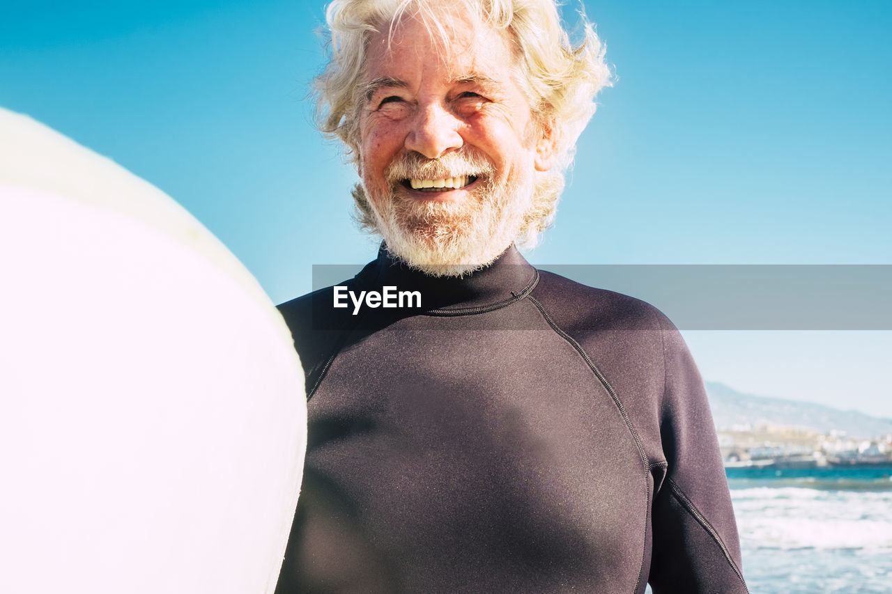 Smiling senior man standing with surfboard at beach on sunny day