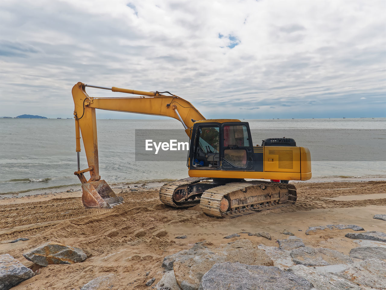 Yellow excavator working on the beach against sea water and cloudy sky