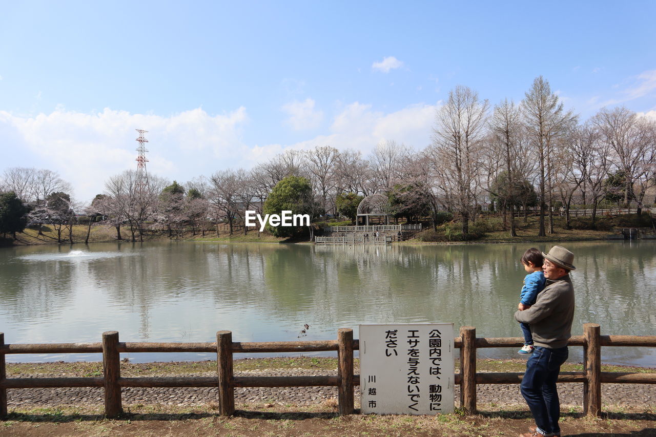 MAN STANDING ON RAILING BY LAKE AGAINST SKY