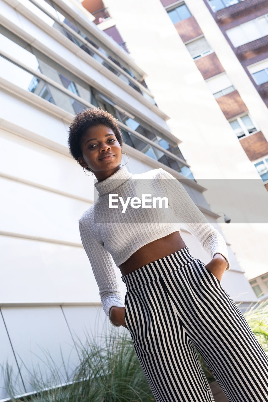 From below stylish self assured african american woman with hands on the pocket looking at camera while standing near modern apartment buildings on sunny day in city
