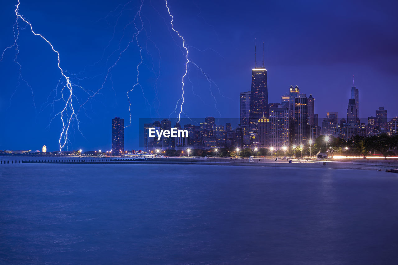 Forked lightning over city by river against sky at night