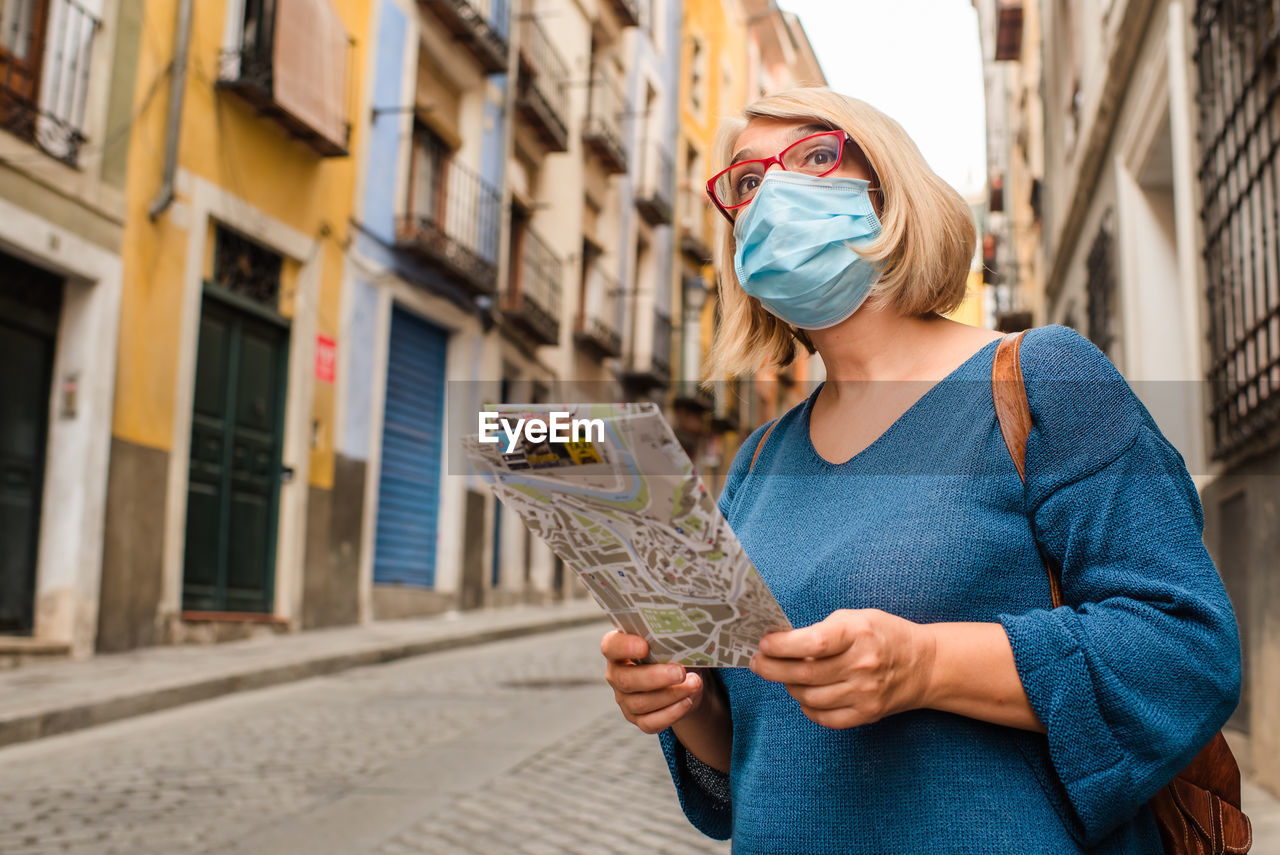 Low angle of female traveler in medical mask for coronavirus prevention holding touristic map and checking route while standing on narrow paved street with old buildings in cuenca town in spain