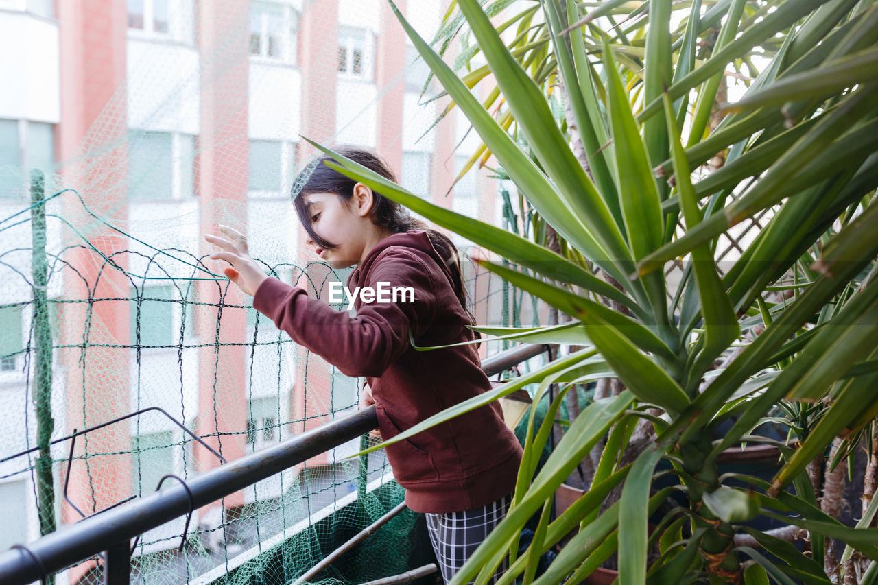 Side view of young girl standing against plants watching outside