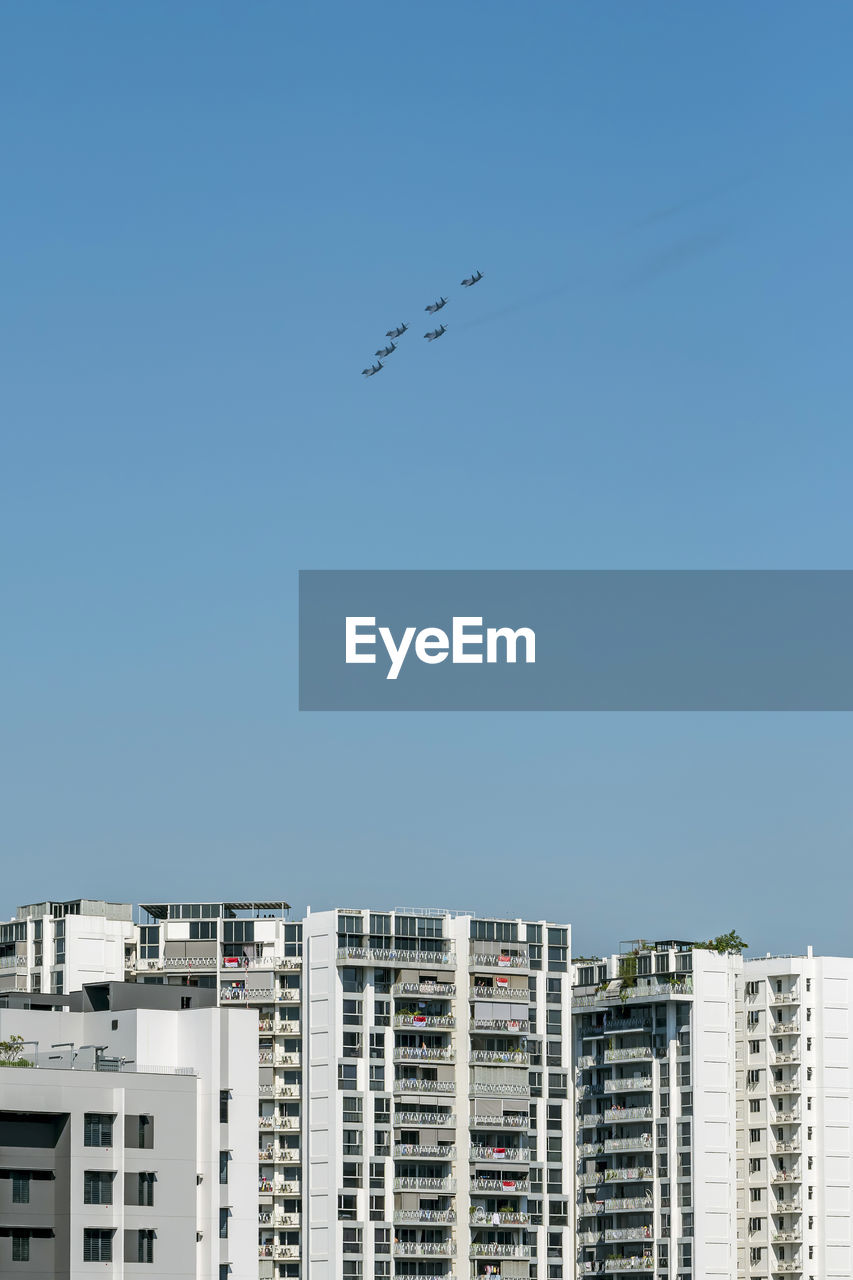 Low angle view of buildings against clear blue sky with airplanes flying overhead