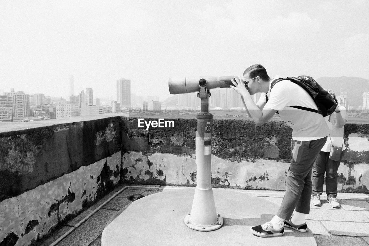 Side view of man standing against sky in city with binoculars