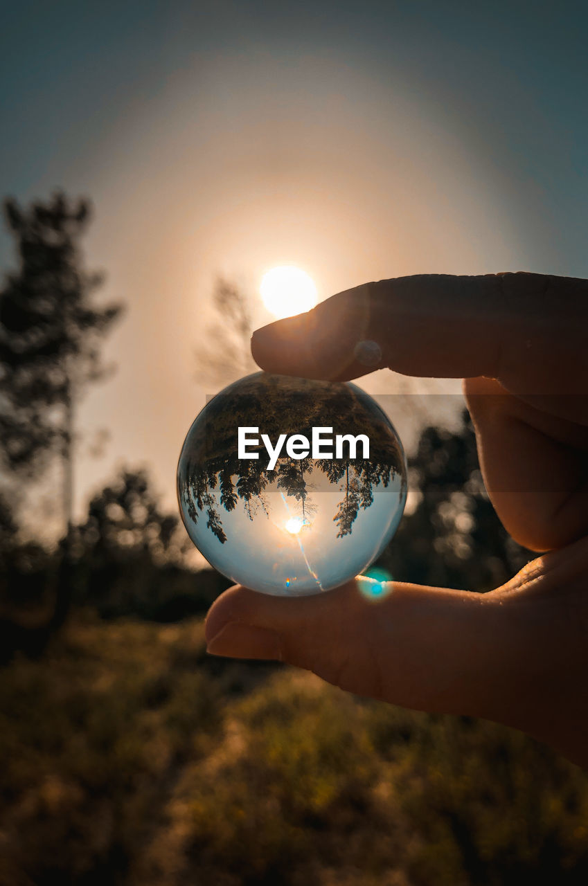 Close-up of hand holding crystal ball against sky during sunset