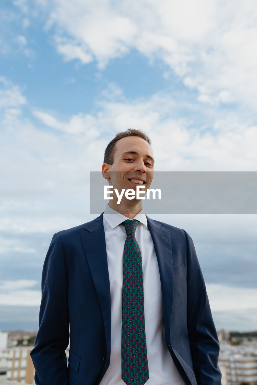 Young businessman in a suit and tie looking at camera and smiling while standing  with a cloudy sky