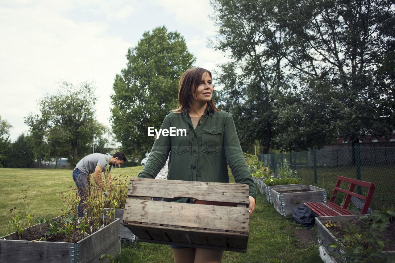 Mid adult woman carrying crate while man planting in background at urban garden