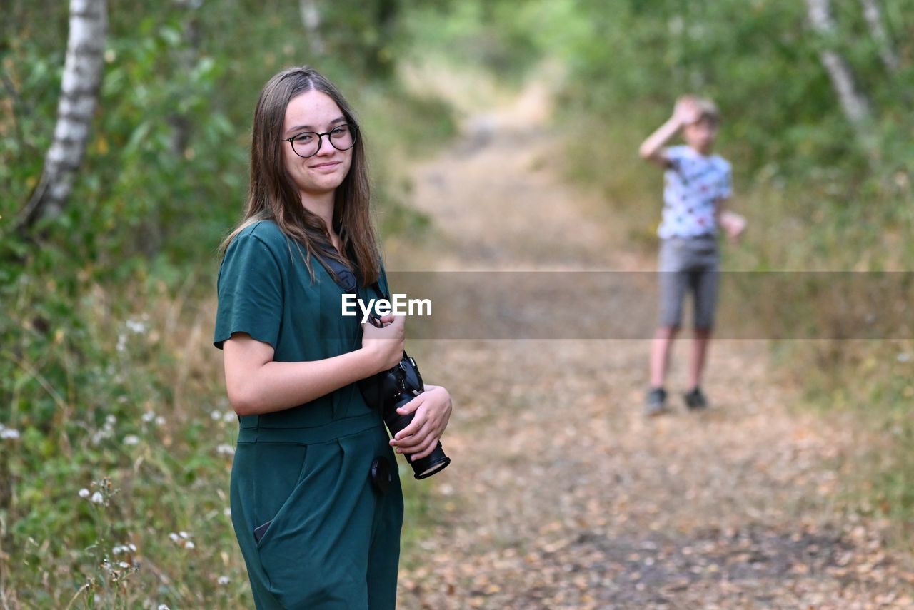 Full length of young woman using camera while standing on field. her brother is behind.