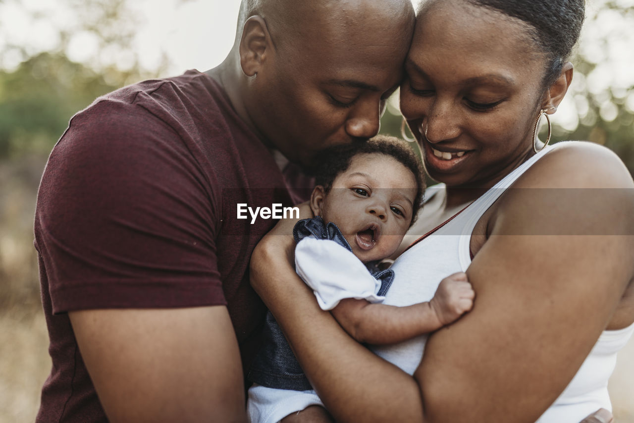 Close up happy mother and father cuddling infant girl backlit field