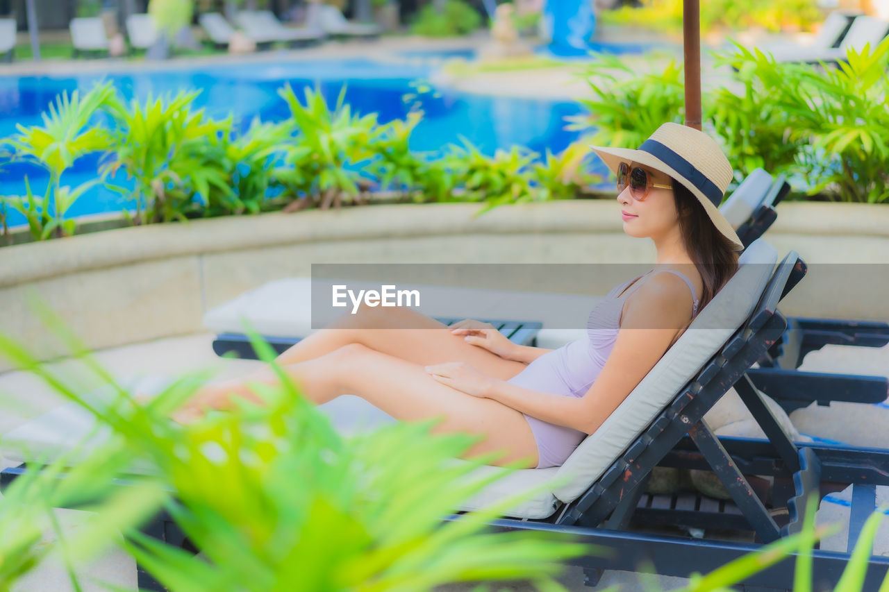 SIDE VIEW OF WOMAN SITTING ON CHAIR AT SWIMMING POOL