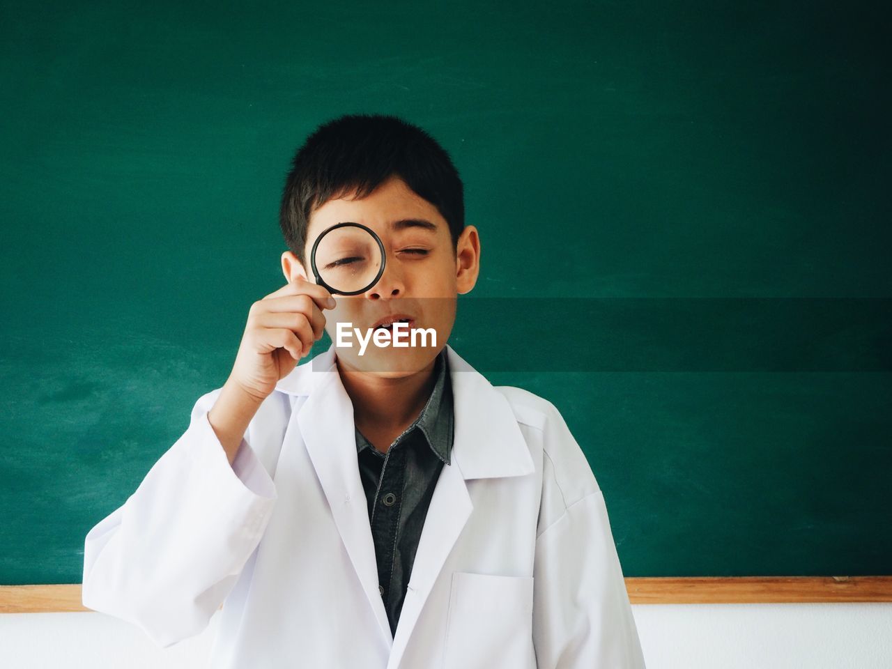 Portrait of boy looking through magnifying glass against blackboard