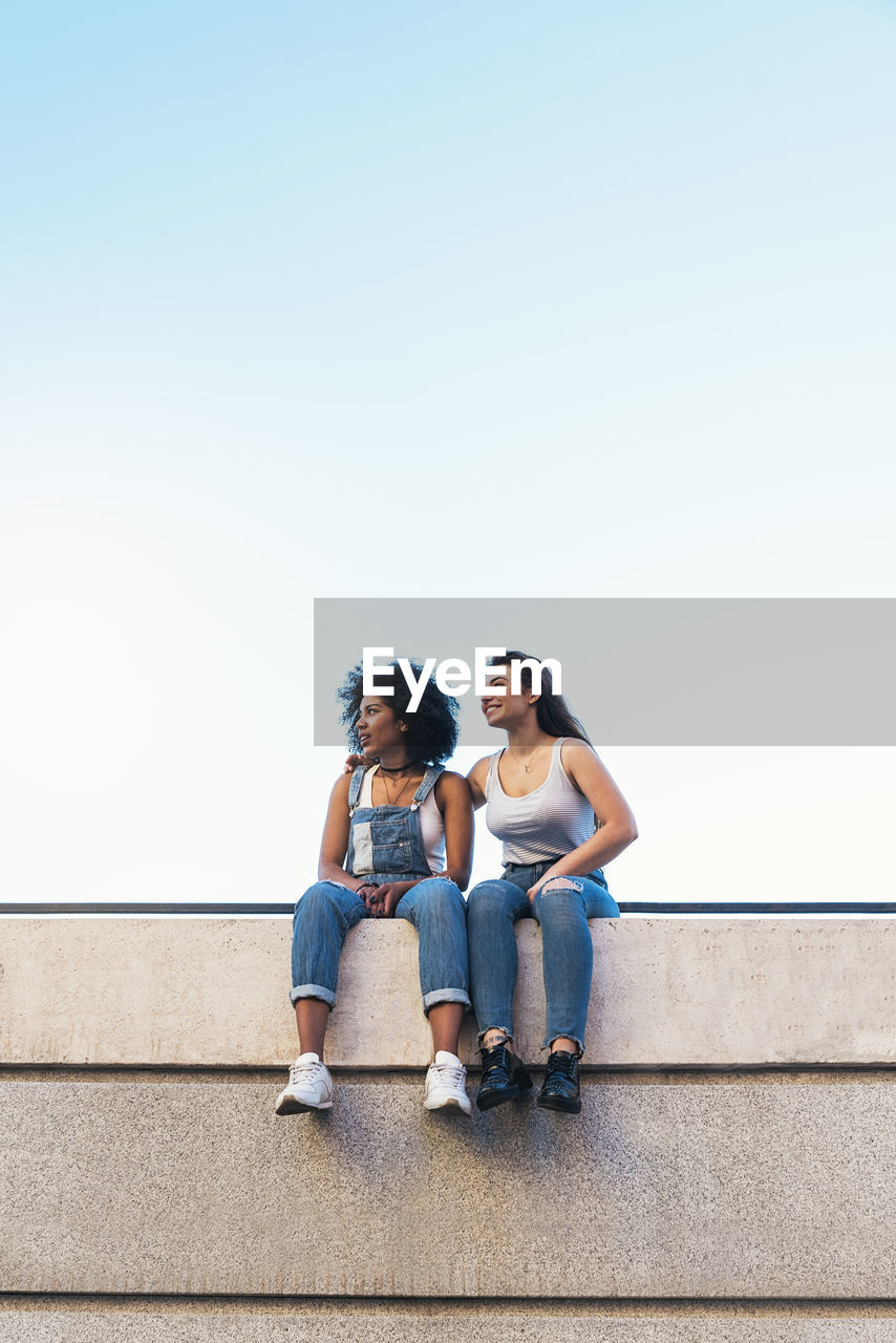 Female friends sitting on retaining wall against clear sky