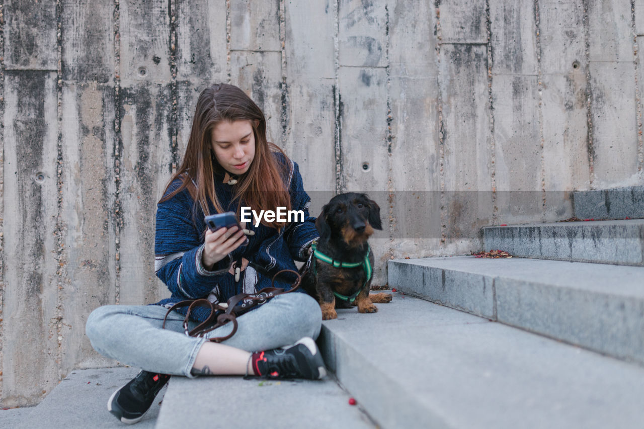 Young adult woman looking her smartphone stairs sit next to her dog