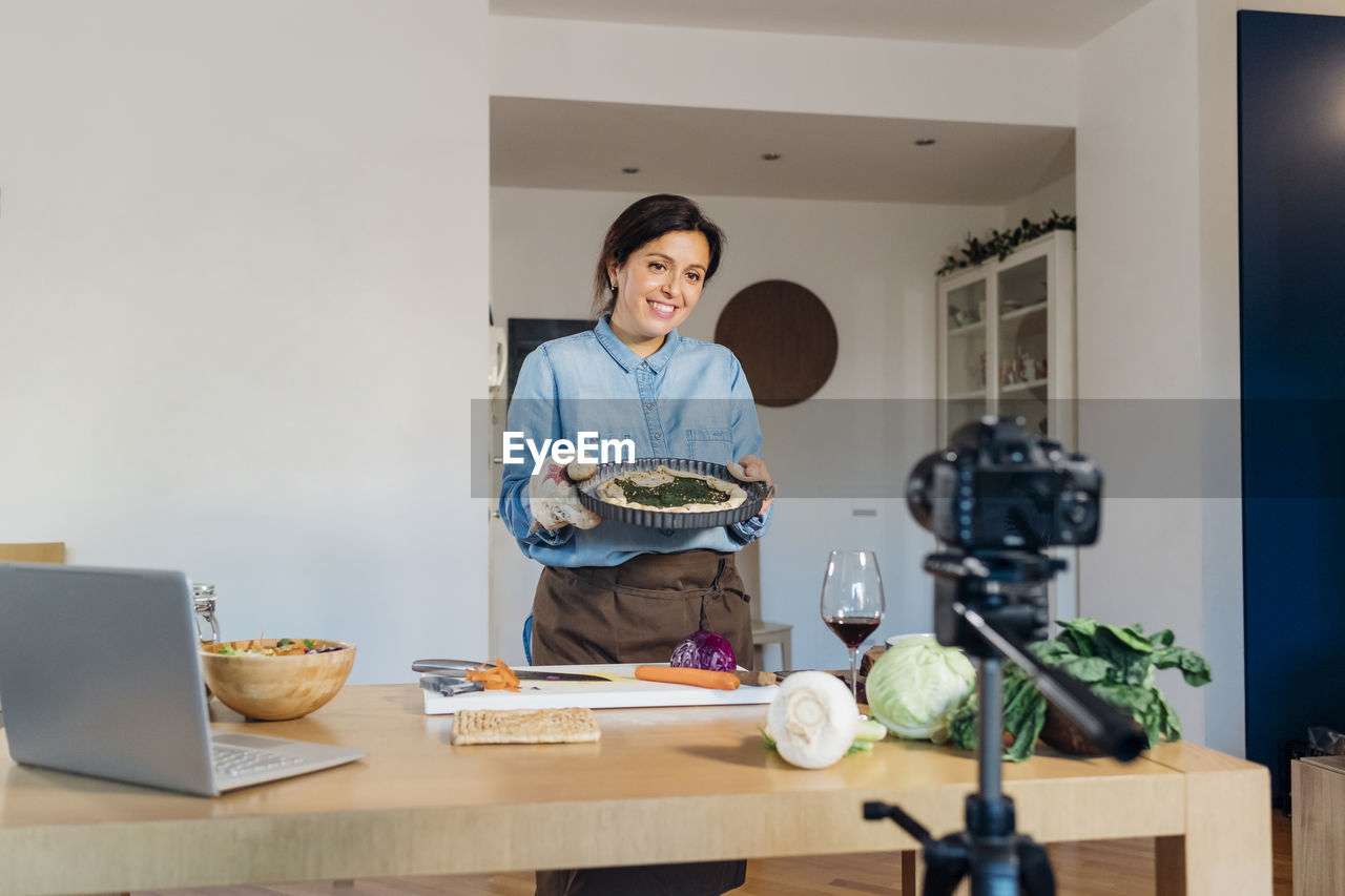 Smiling vlogger holding food plate filming through camera at home