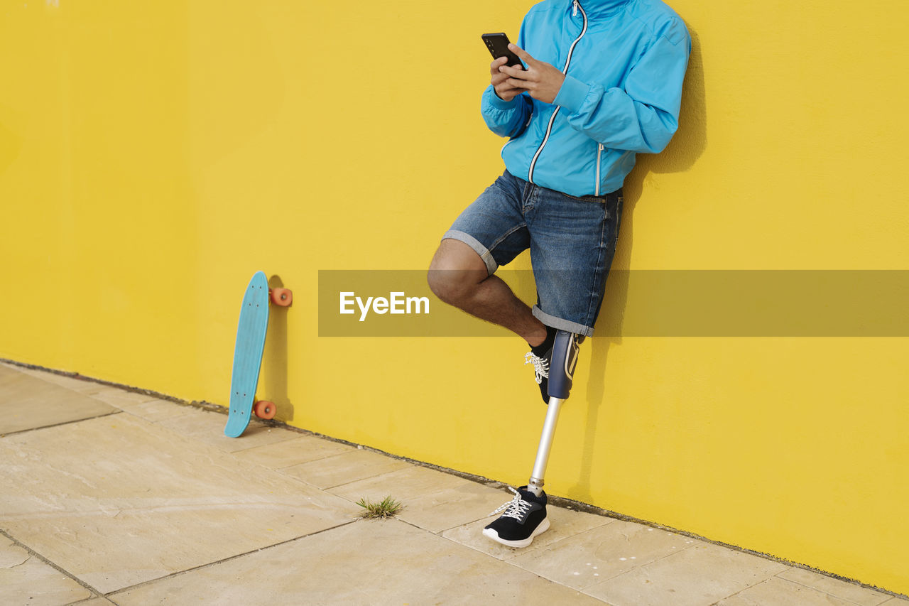 Young disabled athlete using mobile phone while standing against yellow wall