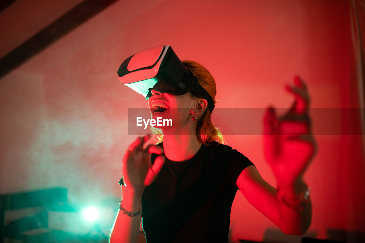Happy woman wearing t shirt while using vr goggles and standing in studio with red neon lights