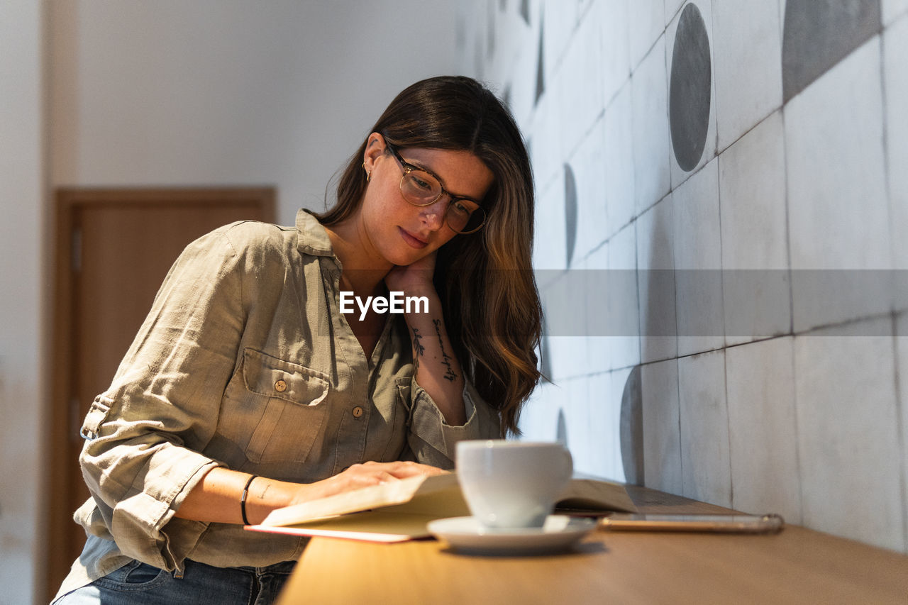 Young wistful female in casual apparel with textbook and cup of hot drink looking away in cafeteria