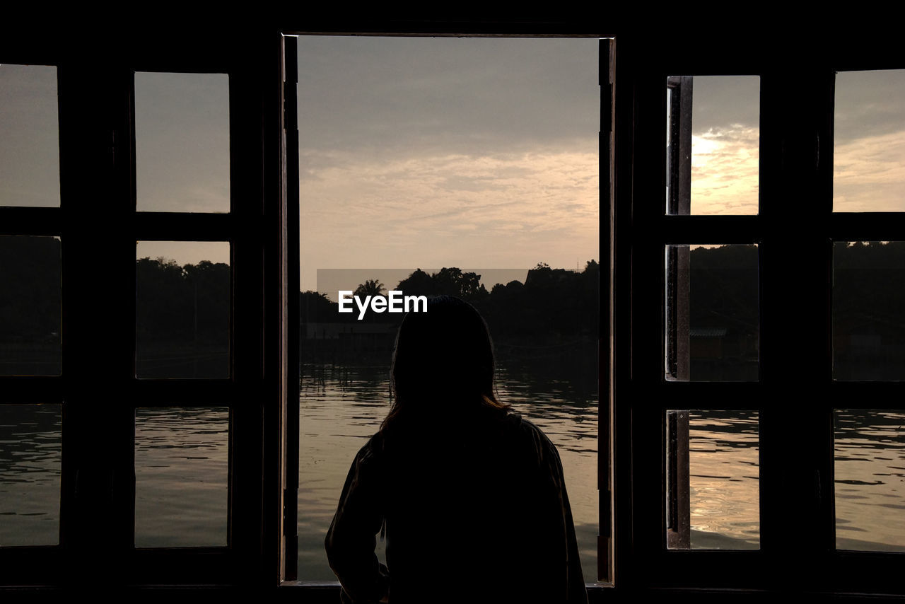 Woman looking at lake through window during sunset