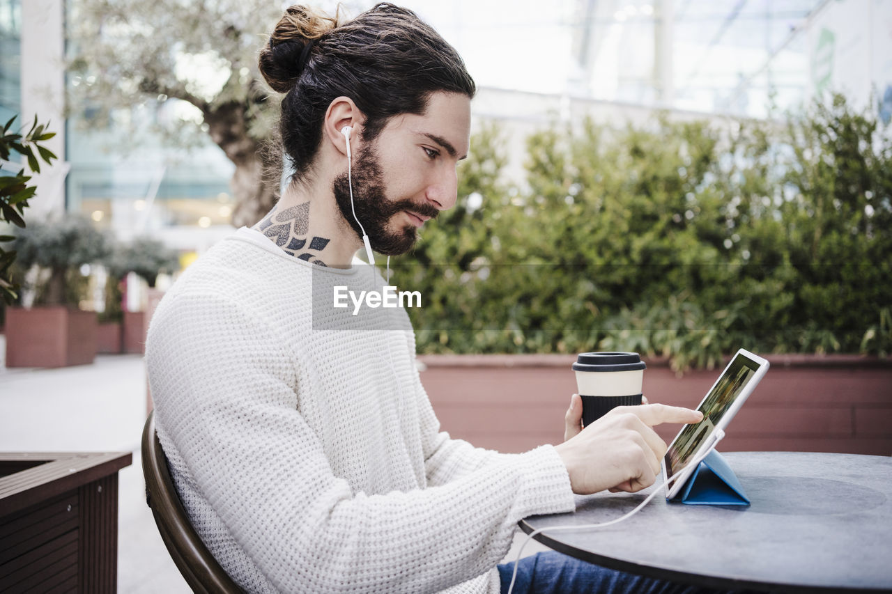 Male hipster with disposable cup touching digital tablet during video call at sidewalk cafe