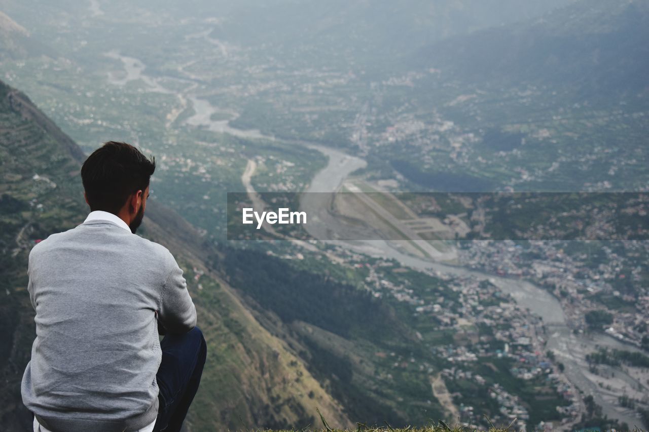 Rear view of man sitting on cliff looking at townscape