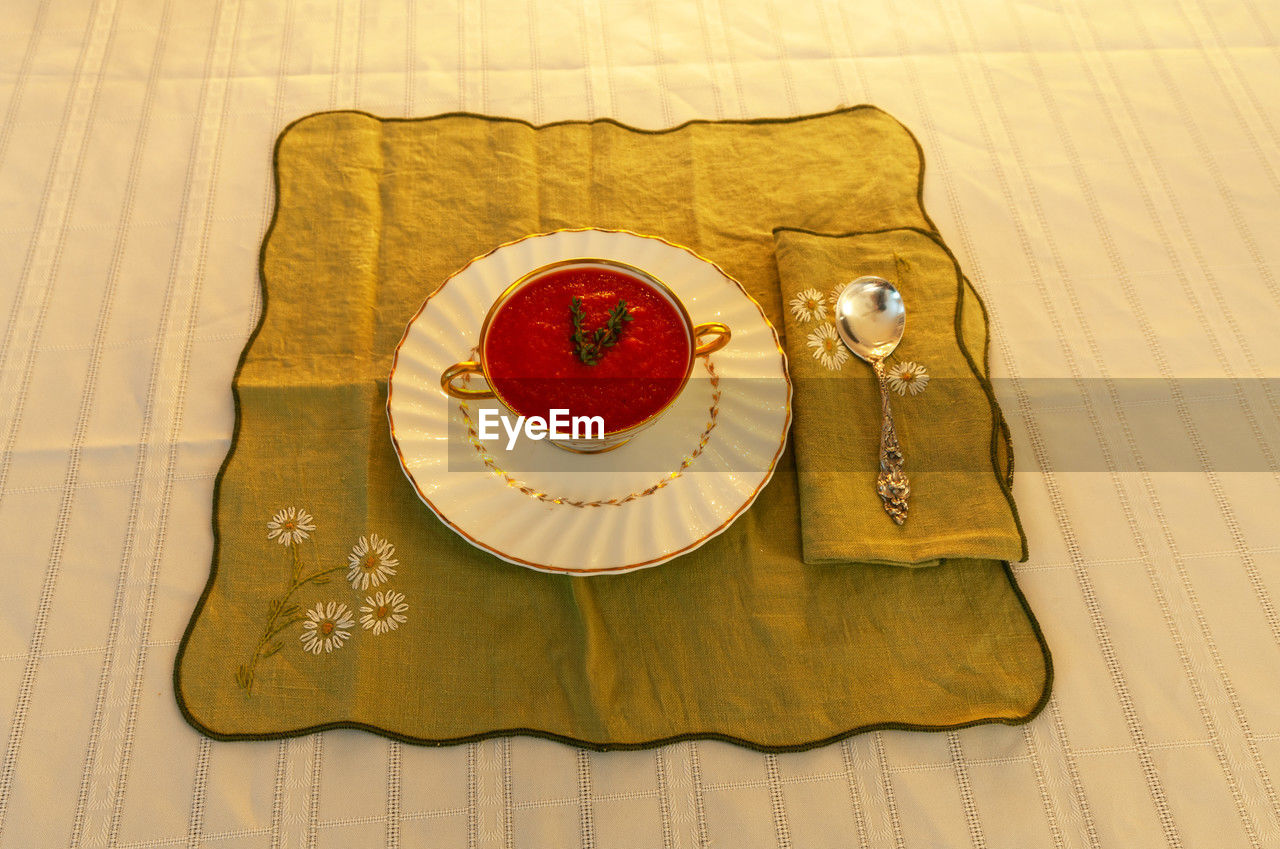 Tomato soup in a white and gold bowl on an embroidered green placemat.