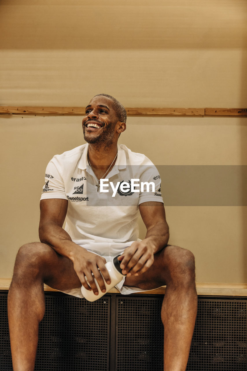 Happy male athlete holding water bottle sitting at sports court