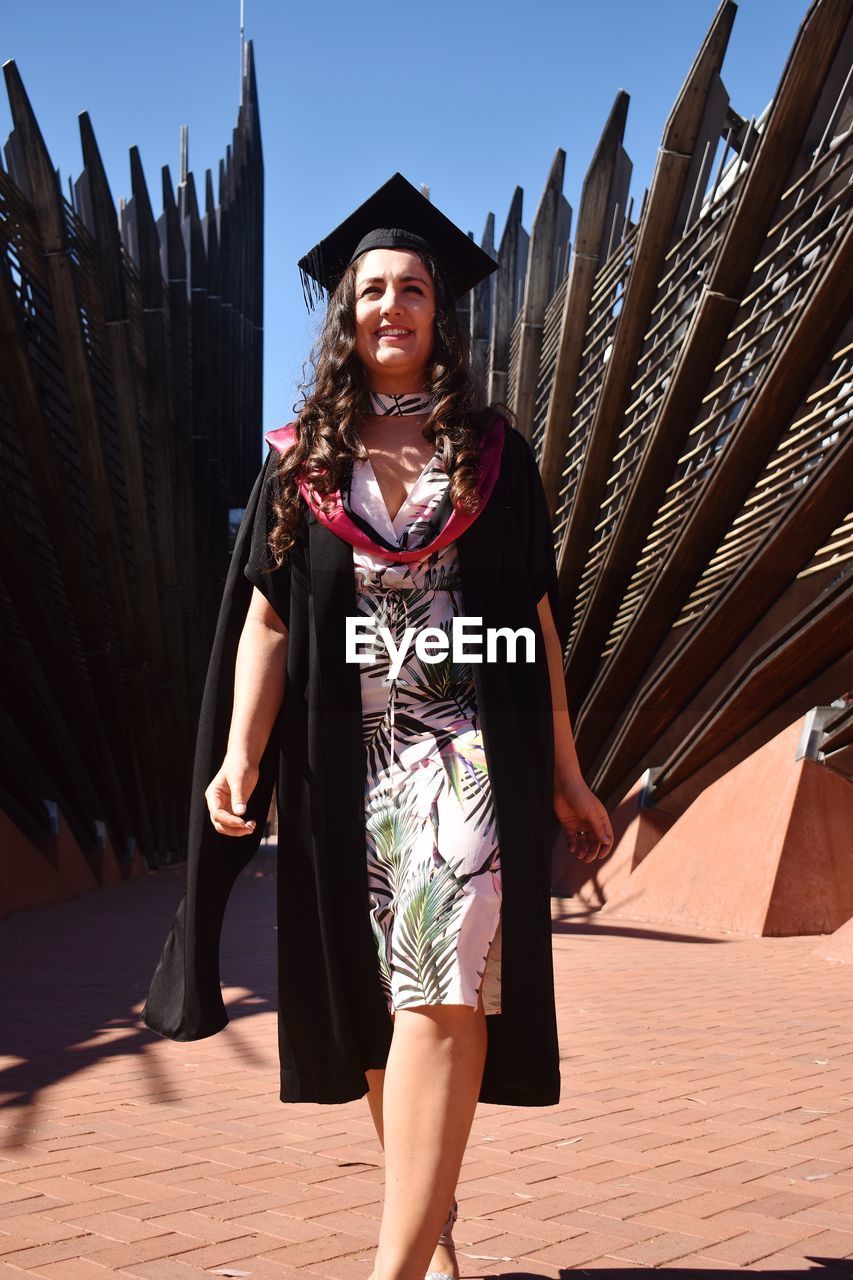 Portrait of smiling young woman standing against building in graduation robes