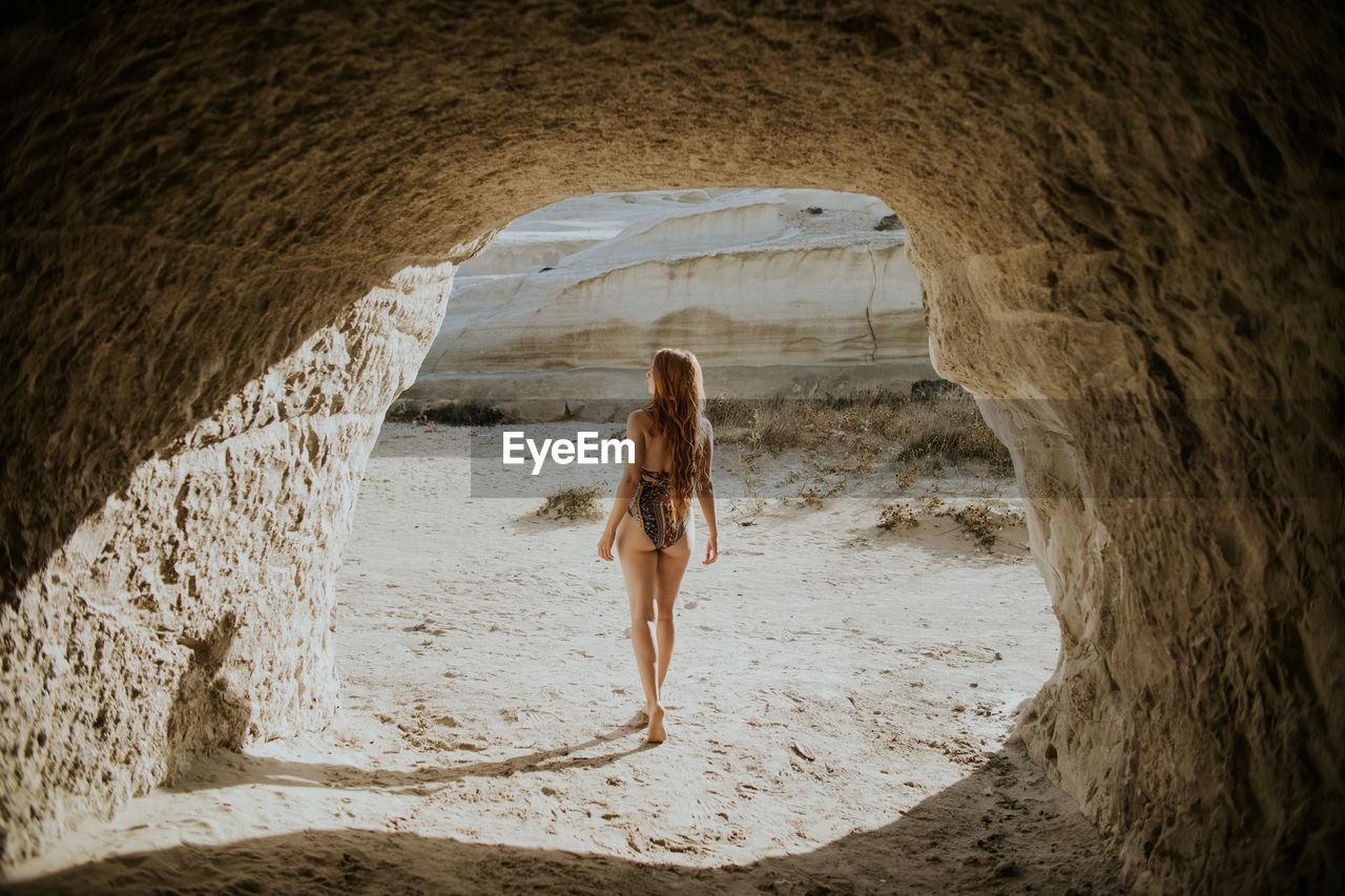 Back view unrecognizable fit female traveler wearing stylish swimsuit walking near stony natural cave in sarakiniko greece on sunny hot day