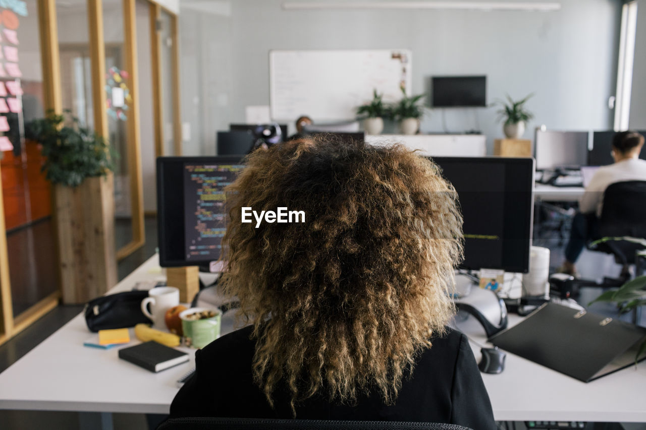 Rear view of female entrepreneur with curly hair sitting and working at desk in office