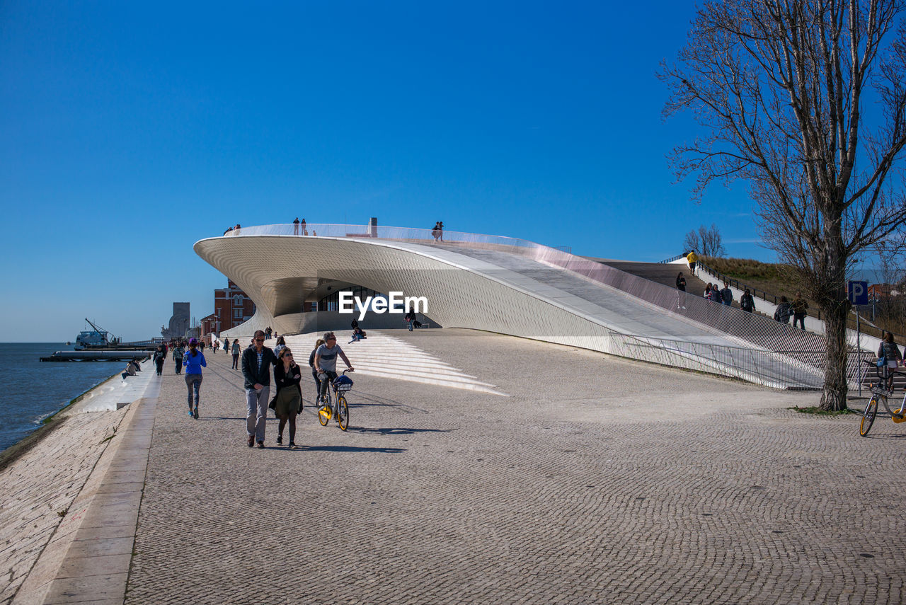 PEOPLE WALKING ON SIDEWALK AGAINST CLEAR BLUE SKY