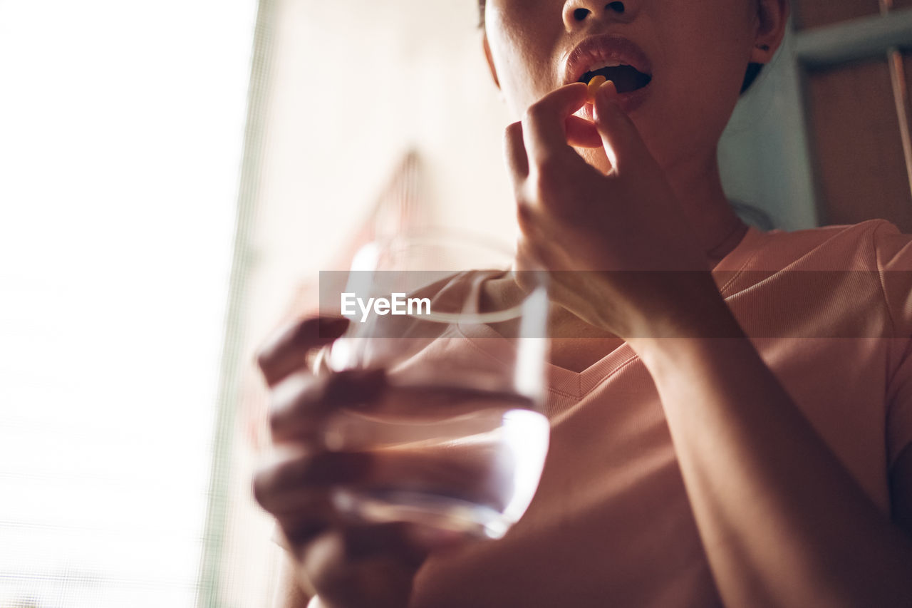 Crop unrecognizable female with glass of fresh water taking yellow vitamin pill while representing healthy lifestyle concept