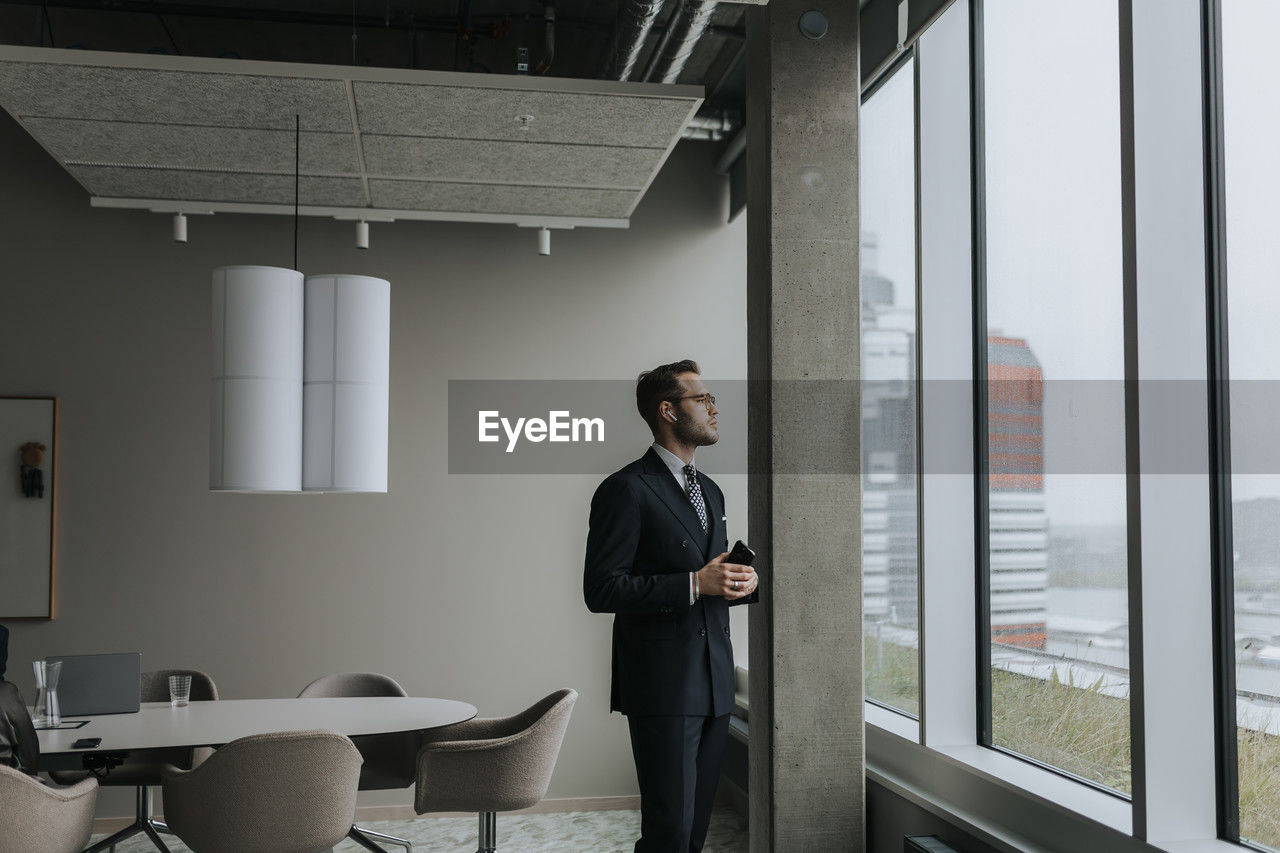 Thoughtful businessman with smart phone looking out through window while standing in board room