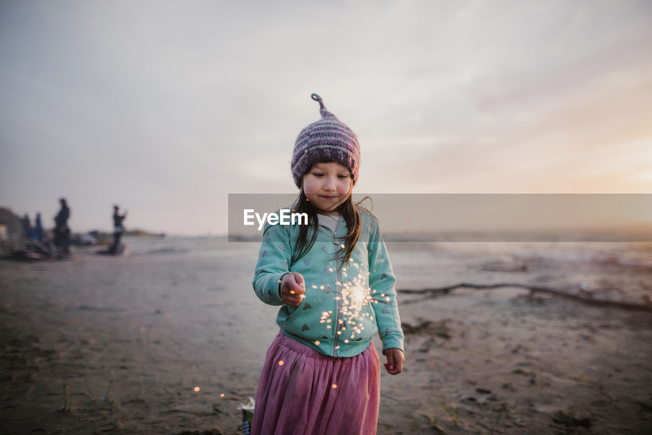 Girl wearing knit hat while holding illuminated sparkler against sky at beach