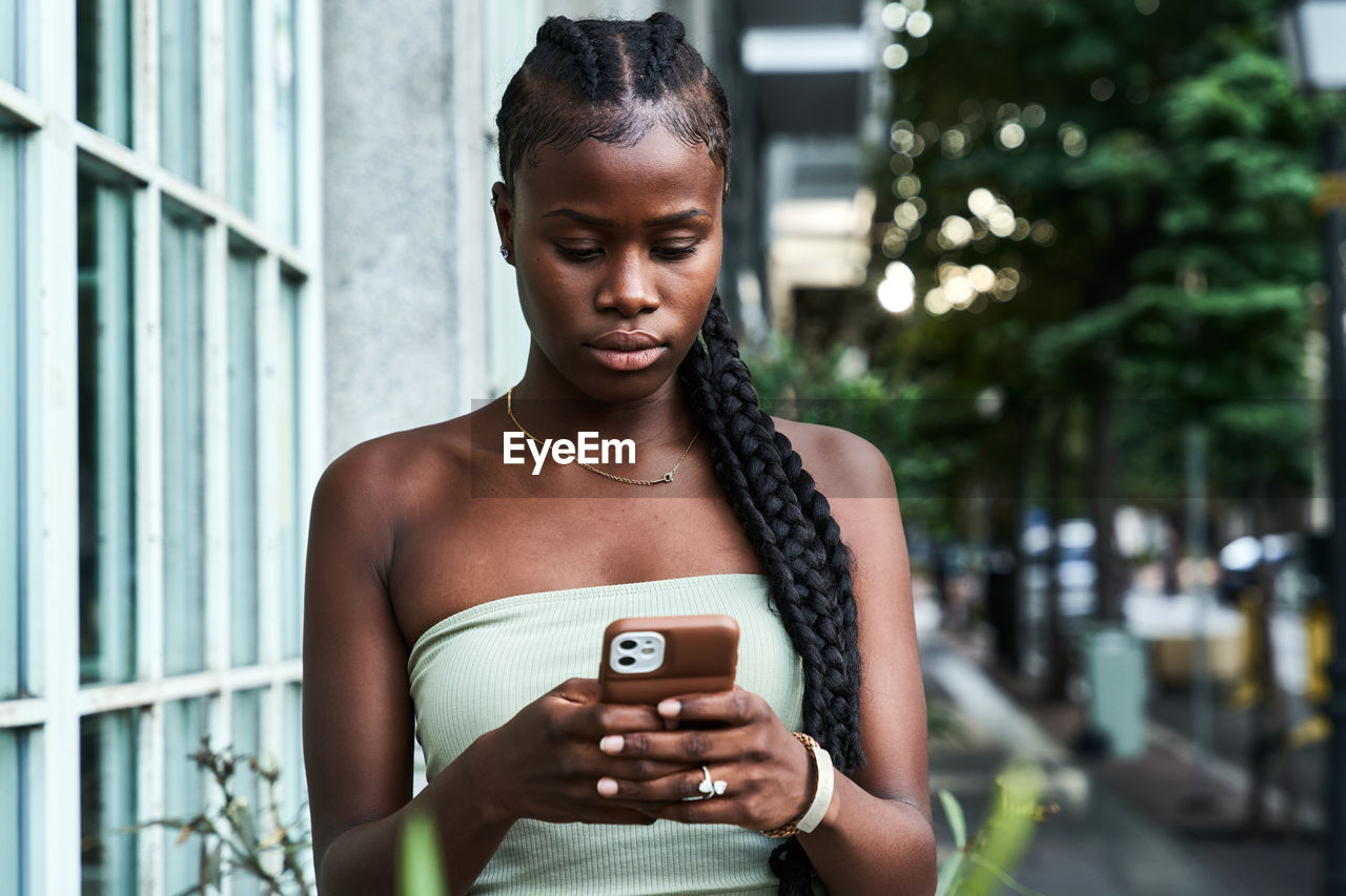 Young african american woman with long braids wearing stylish tube top and browsing smartphone on city street